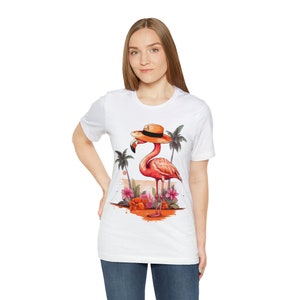 May include: White t-shirt with a pink flamingo wearing a brown hat and standing in front of palm trees and flowers.