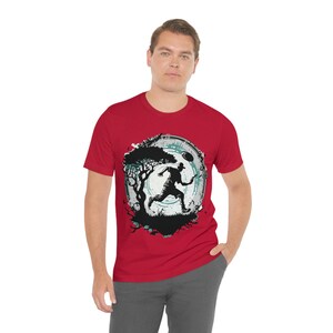 May include: A red t-shirt with a graphic of a person running with a frisbee in front of a moon and a tree. The graphic is in black and white with a teal blue outline.