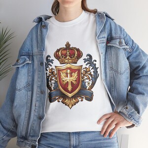 May include: White t-shirt featuring a detailed crest design. The crest includes a red and gold shield with a cross, topped with a red and gold crown. The design is framed by ornate blue and gold flourishes.