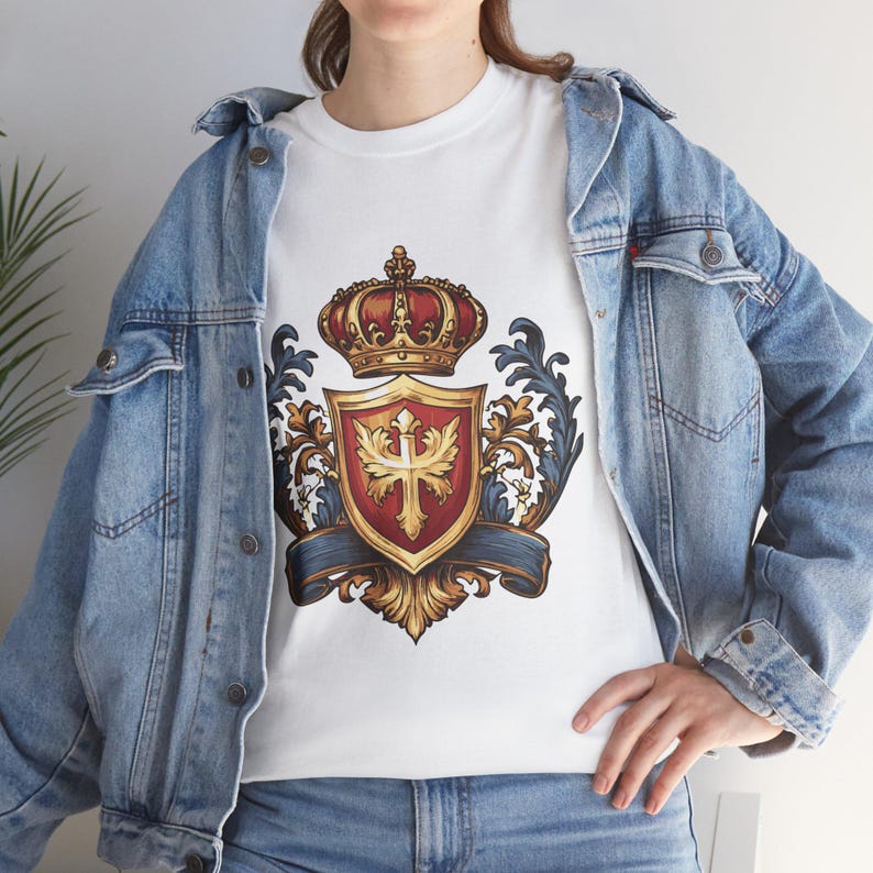 May include: White t-shirt featuring a detailed crest design. The crest includes a red and gold shield with a cross, topped with a red and gold crown. The design is framed by ornate blue and gold flourishes.