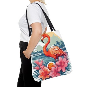 May include: A white tote bag with a pink flamingo standing in a tropical scene with pink flowers. The flamingo is in front of a blue body of water and a sunset.