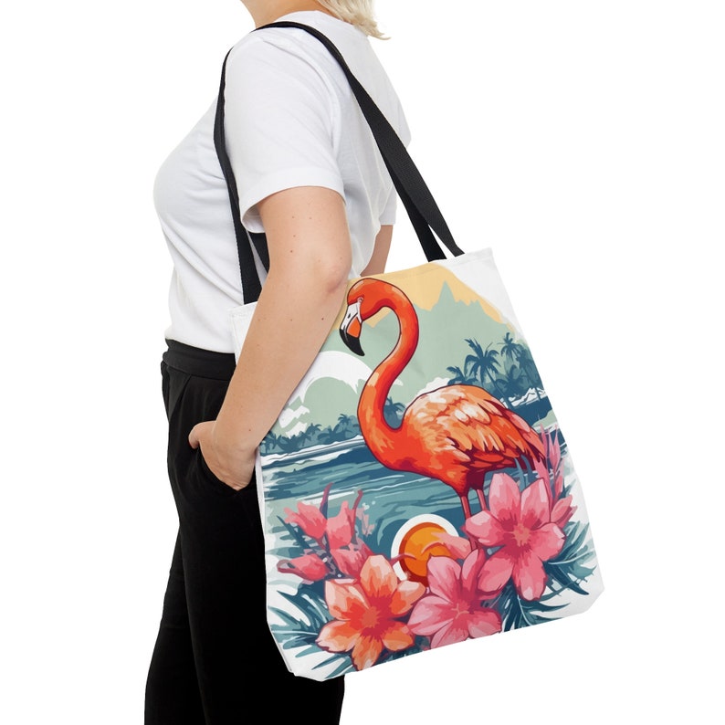 May include: A white tote bag with a pink flamingo standing in a tropical scene with pink flowers. The flamingo is in front of a blue body of water and a sunset.
