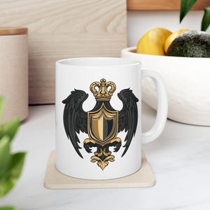 May include: White ceramic mug featuring a black and gold emblem. The emblem includes a crown, shield, and stylized wings. The mug has a curved handle and is sitting on a beige coaster. The design is centered on the mug.