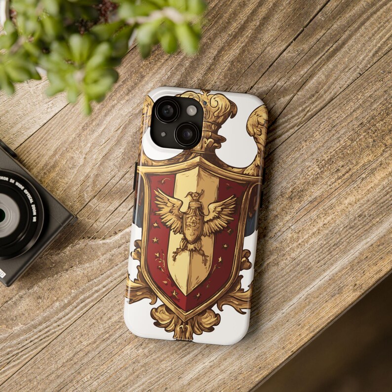 May include: A white phone case featuring a detailed heraldic crest design. The crest is primarily gold, red, and white, with a central eagle emblem. The case has a glossy finish and is designed to fit a smartphone.