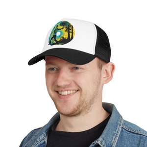 May include: A black and white trucker hat with a white front panel featuring a graphic of a person walking through a forest with a yellow sun and blue sky in the background. The hat has a black mesh back panel.