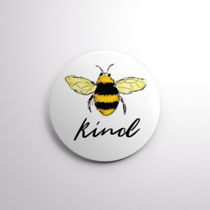 May include: A white circular badge featuring a watercolour-style bee in yellow and black. The word "kind" is written in black script below the bee. The badge has a glossy finish.