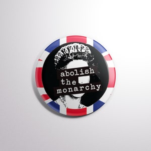 May include: A black and white button featuring a portrait of a woman wearing a crown. The woman's eyes are covered with a black bar that reads "abolish the monarchy". The button is surrounded by a red, white and blue Union Jack flag.