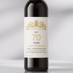 May include: A dark glass wine bottle with a white rectangular label featuring a gold ornate flourish at the top. The text reads VINTAGE EST 70 YEARS Aged to Perfection Happy 70th Birthday Jeanne followed by a list of names including Dave, Julie, Stu, Jo, Nik, Klaire, Jake, Toby, Dylan, Cyena, Alice & Scarlett.