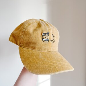 May include: A mustard yellow baseball cap with an embroidered design of a honey pot with a dipper and a crescent moon.