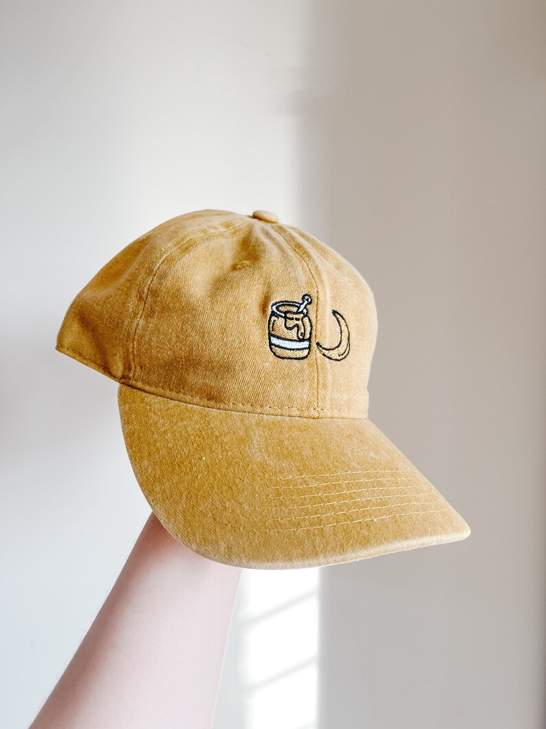 May include: A mustard yellow baseball cap with an embroidered design of a honey pot with a dipper and a crescent moon.