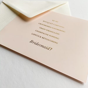 Puede incluir: Una tarjeta rosa con letras doradas que dice "Be my Bridesmaid?" con una lista de tareas: "Bouquet Carrying, Groomsmen Gathering, Train Wrangling, Champagne Popping, Lipstick Retouching."