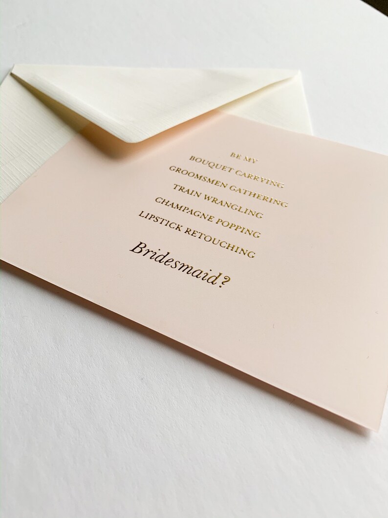 Puede incluir: Una tarjeta rosa con letras doradas que dice "Be my Bridesmaid?" con una lista de tareas: "Bouquet Carrying, Groomsmen Gathering, Train Wrangling, Champagne Popping, Lipstick Retouching."