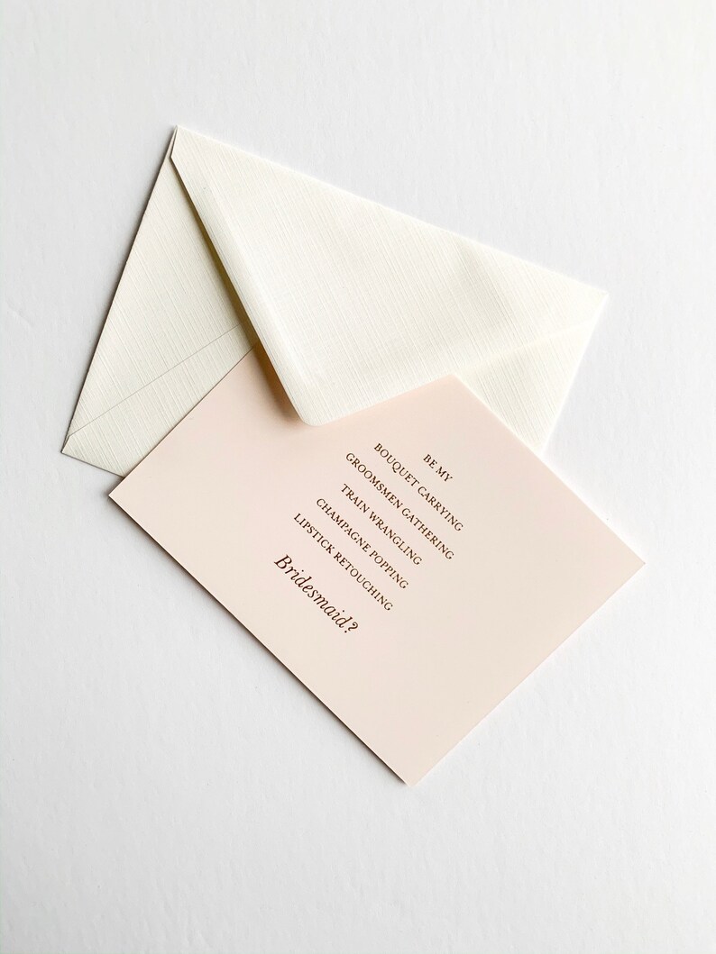 Puede incluir: Una tarjeta rosa con el texto "Be My Bridesmaid?" y una lista de tareas: "Bouquet Carrying", "Groomsmen Gathering", "Train Wrangling", "Champagne Popping", "Lipstick Retouching".