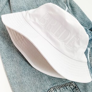 May include: White bucket hat with the word "BRIDE" embroidered in white on the front. The hat is lying on a blue denim jacket.