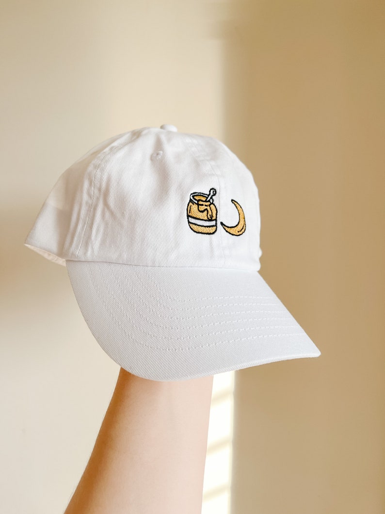 May include: White baseball cap with an embroidered design of a jar of honey and a crescent moon.