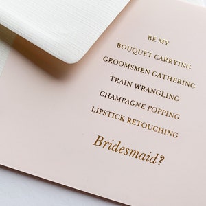 Puede incluir: Una tarjeta rosa con letras doradas que dice "Be my bridesmaid?" con una lista de tareas: "Bouquet carrying", "Groomsmen gathering", "Train wrangling", "Champagne popping" y "Lipstick retouching".