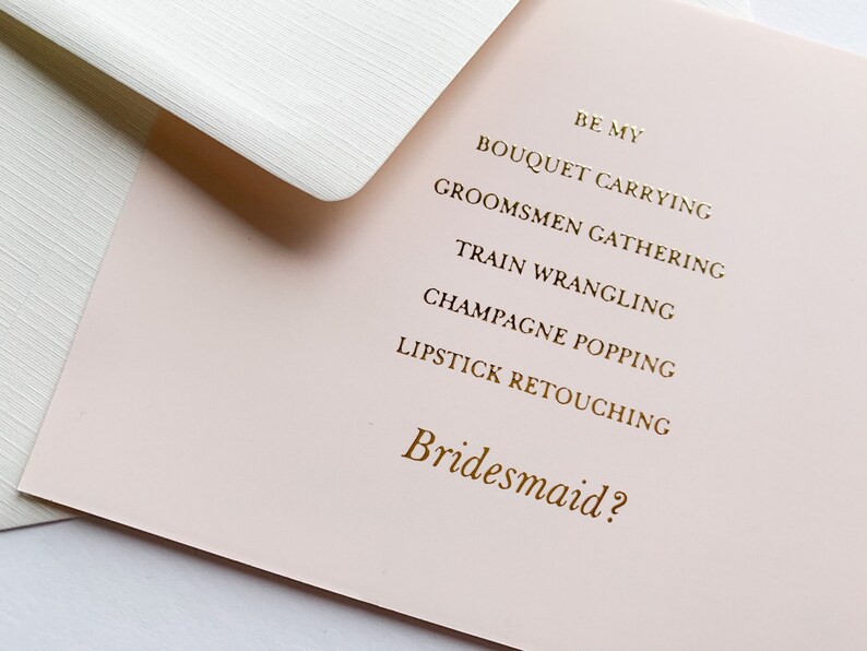 Puede incluir: Una tarjeta rosa con letras doradas que dice "Be my bridesmaid?" con una lista de tareas: "Bouquet carrying", "Groomsmen gathering", "Train wrangling", "Champagne popping" y "Lipstick retouching".