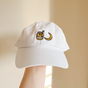 May include: White baseball cap with an embroidered design of a jar of honey and a crescent moon.