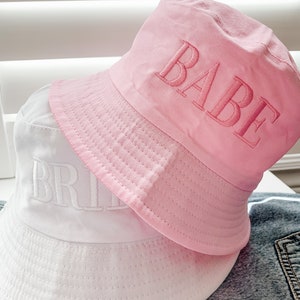 May include: Two bucket hats, one white with the word "BRIDE" embroidered in white, and one pink with the word "BABE" embroidered in pink.