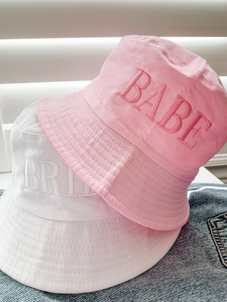 May include: Two bucket hats, one white with the word "BRIDE" embroidered in white, and one pink with the word "BABE" embroidered in pink.