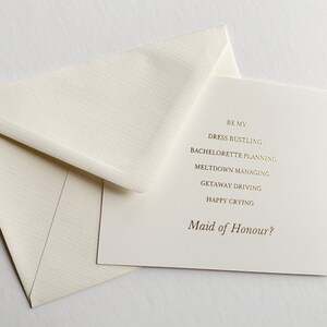 Puede incluir: Una tarjeta blanca con letras doradas que dice "Be my Dress Bustling, Bachelorette Planning, Meltdown Managing, Getaway Driving, Happy Crying, Maid of Honour?"