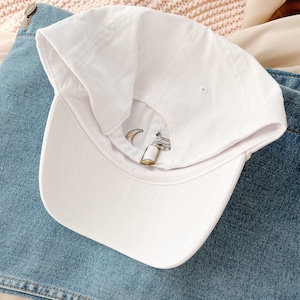 May include: White baseball cap with a crescent moon and star embroidery. The cap is on a blue denim background.