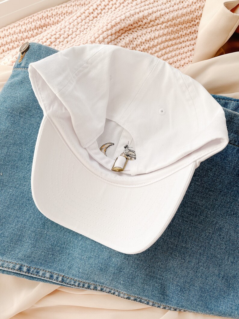May include: White baseball cap with a crescent moon and star embroidery. The cap is on a blue denim background.