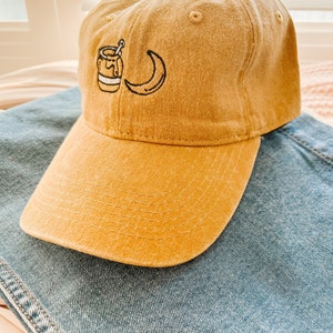 May include: A mustard yellow baseball cap with a small embroidered design of a jar of honey and a crescent moon.