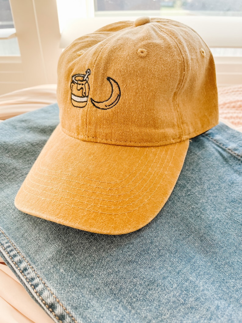 May include: A mustard yellow baseball cap with a small embroidered design of a jar of honey and a crescent moon.