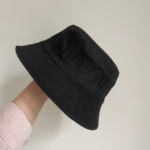 May include: Black bucket hat with the word "GROOM" embroidered in black on the front.