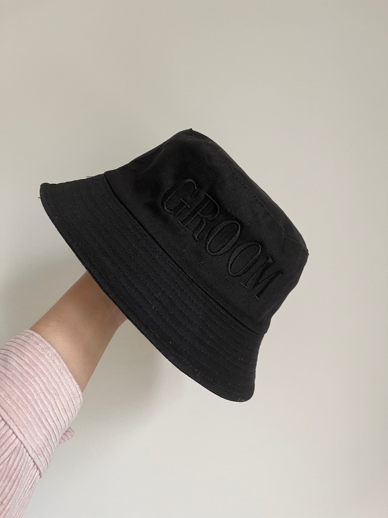 May include: Black bucket hat with the word "GROOM" embroidered in black on the front.