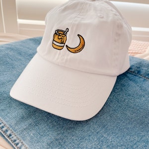 May include: White baseball cap with an embroidered design of a honey pot and a crescent moon.