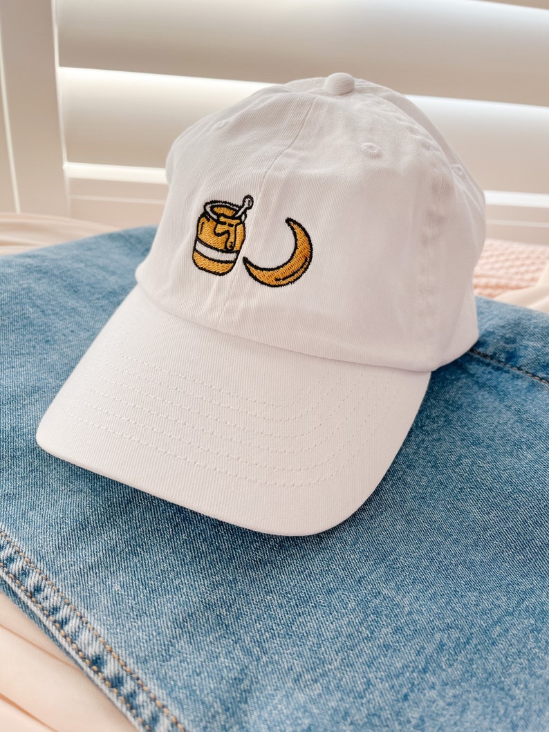 May include: White baseball cap with an embroidered design of a honey pot and a crescent moon.