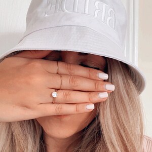 May include: A white bucket hat with the word "BRIDE" embroidered in white on the front. The hat is worn by a person with long blonde hair, and the person is covering their face with their hand.