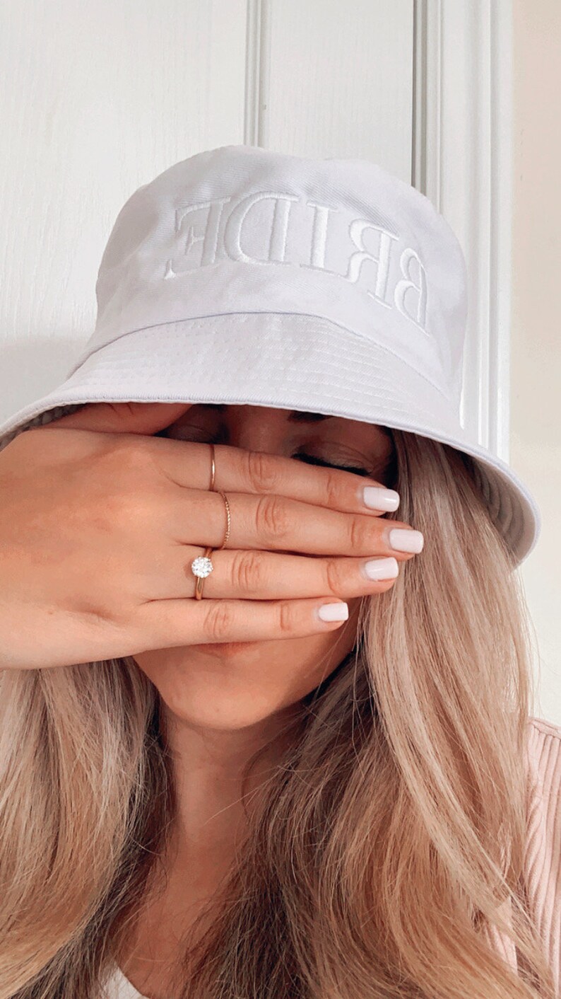 May include: A white bucket hat with the word "BRIDE" embroidered in white on the front. The hat is worn by a person with long blonde hair, and the person is covering their face with their hand.