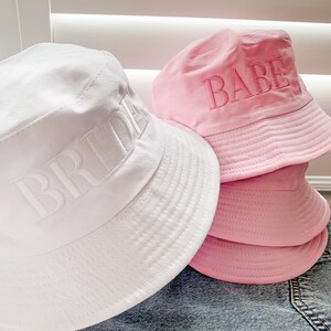 May include: Two white and pink bucket hats with the words "Bride" and "Babe" embroidered on them.