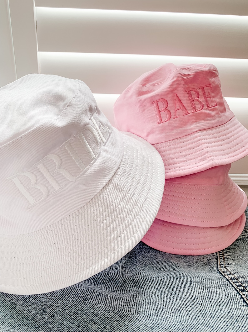 May include: Two white and pink bucket hats with the words "Bride" and "Babe" embroidered on them.