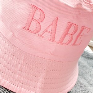 May include: A pink bucket hat with the word "BABE" embroidered on the front.