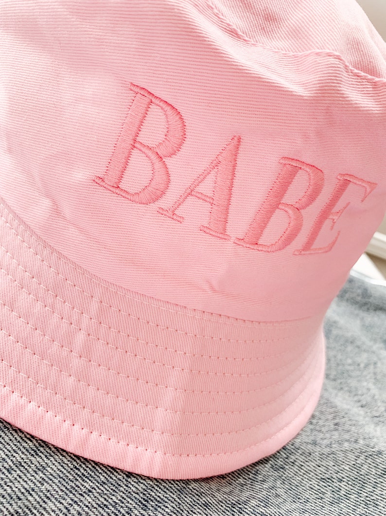 May include: A pink bucket hat with the word "BABE" embroidered on the front.