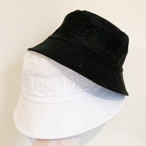 May include: Two bucket hats, one black and one white, with the words "Groom" and "Bride" embroidered on them.