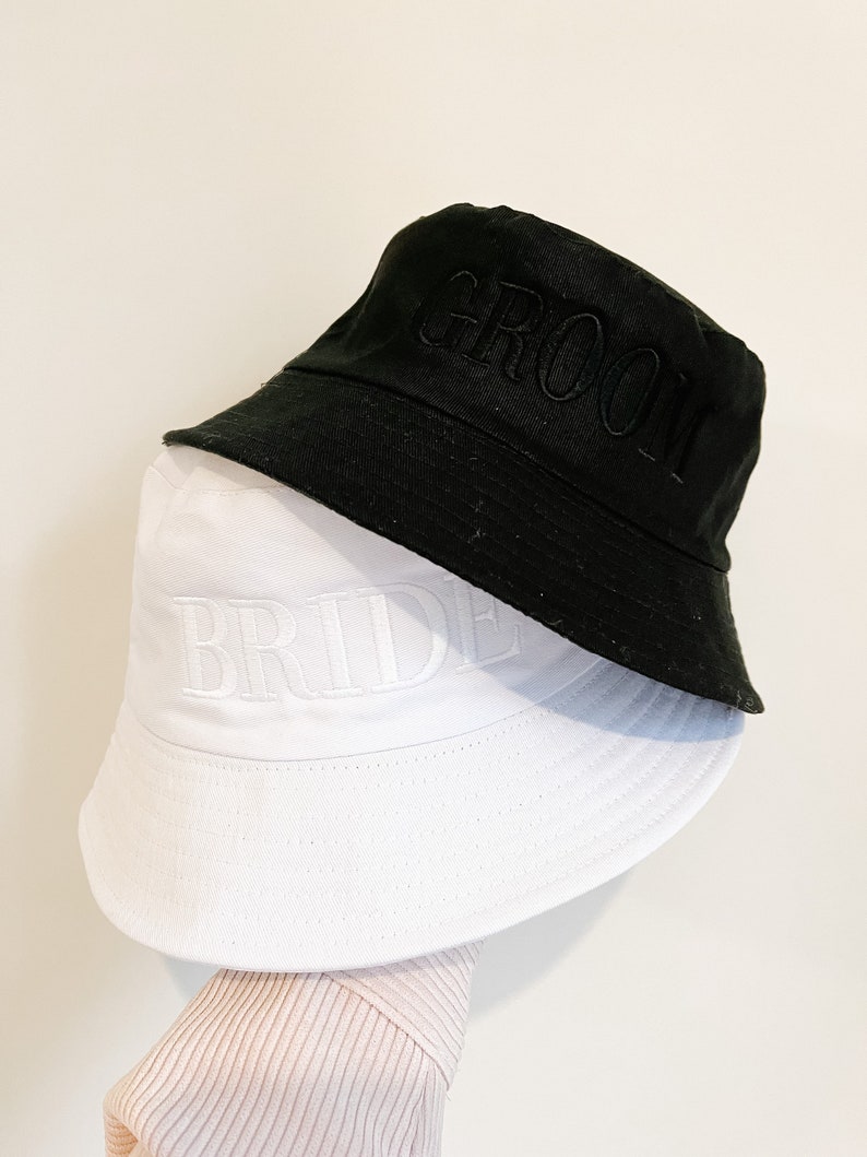 May include: Two bucket hats, one black and one white, with the words "Groom" and "Bride" embroidered on them.
