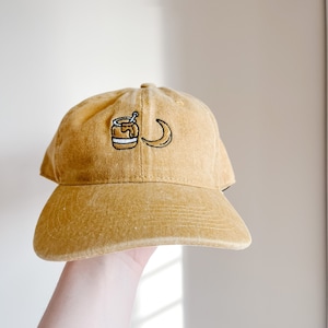 May include: A mustard yellow baseball cap with an embroidered design of a jar of honey and a crescent moon.