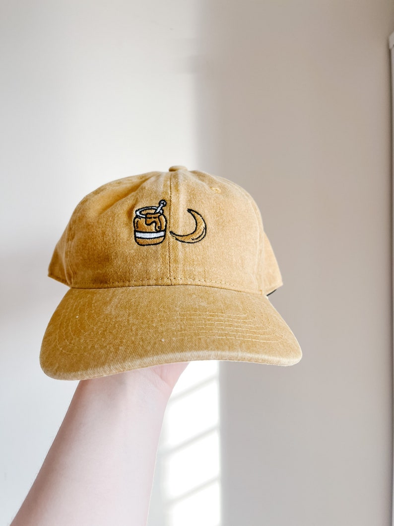 May include: A mustard yellow baseball cap with an embroidered design of a jar of honey and a crescent moon.