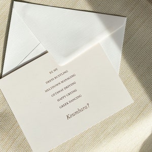 Puede incluir: Una tarjeta blanca con el texto "Be my Dress Bustling Meltdown Managing Getaway Driving Happy Crying Greek Dancing Koumbara?" impreso en tinta marr&oacute;n.