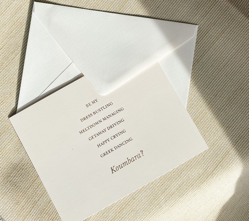 Puede incluir: Una tarjeta blanca con el texto "Be my Dress Bustling Meltdown Managing Getaway Driving Happy Crying Greek Dancing Koumbara?" impreso en tinta marr&oacute;n.