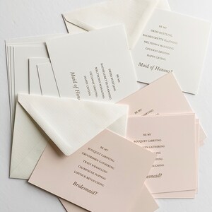 Puede incluir: Un conjunto de tarjetas con el texto "Be My Bridesmaid?" y "Be My Maid of Honor?" impreso en ellas. Las tarjetas est&aacute;n sobre una superficie blanca.