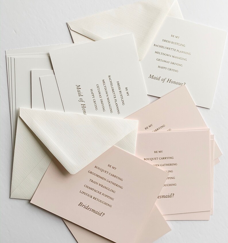Puede incluir: Un conjunto de tarjetas con el texto "Be My Bridesmaid?" y "Be My Maid of Honor?" impreso en ellas. Las tarjetas est&aacute;n sobre una superficie blanca.