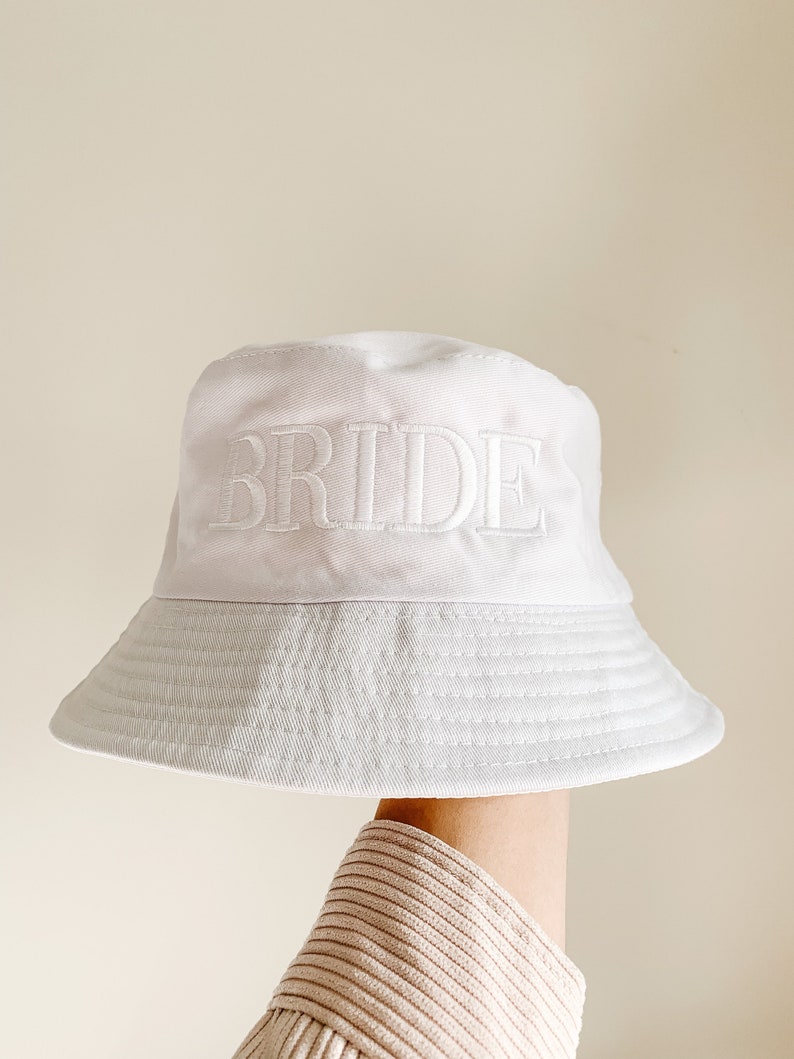 May include: White bucket hat with the word "BRIDE" embroidered in white on the front.