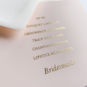 Puede incluir: Una tarjeta rosa con letras doradas que dice "Be my bridesmaid?" con una lista de tareas: "Bouquet carrying", "Groomsmen gathering", "Train wrangling", "Champagne popping" y "Lipstick retouching".