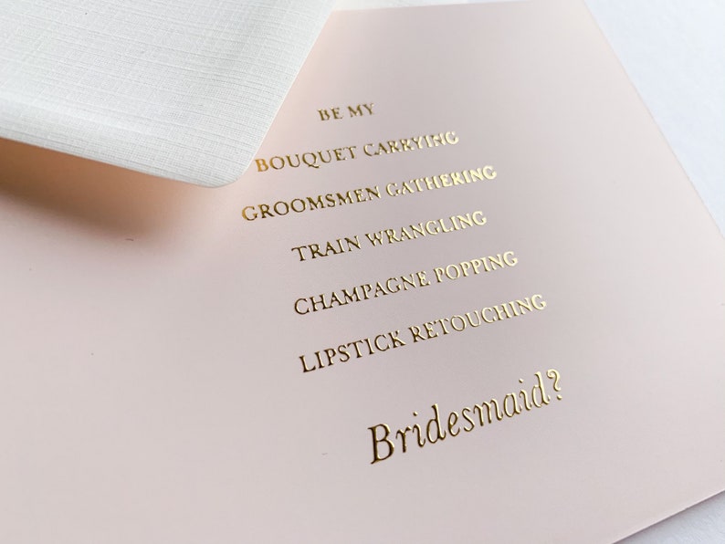 Puede incluir: Una tarjeta rosa con letras doradas que dice "Be my bridesmaid?" con una lista de tareas: "Bouquet carrying", "Groomsmen gathering", "Train wrangling", "Champagne popping" y "Lipstick retouching".